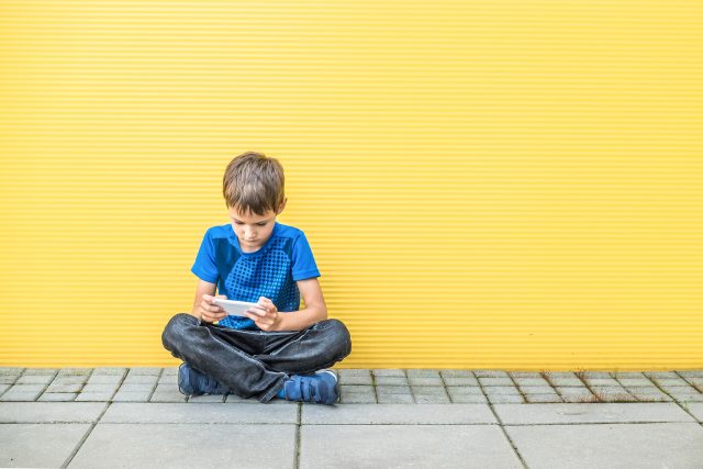 A young boy in a blue shirt sits cross-legged on the ground, looking intently at a mobile phone. He is against a bright yellow wall and grey pavement.