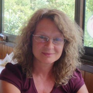 A woman with curly, light brown hair and glasses is seated indoors near large windows, wearing a maroon top and smiling slightly at the camera. Greenery is visible outside the windows.