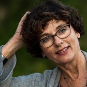 A woman with short curly brown hair and glasses smiles gently while resting one hand on her head, wearing a grey top, outdoors with a blurred green background.