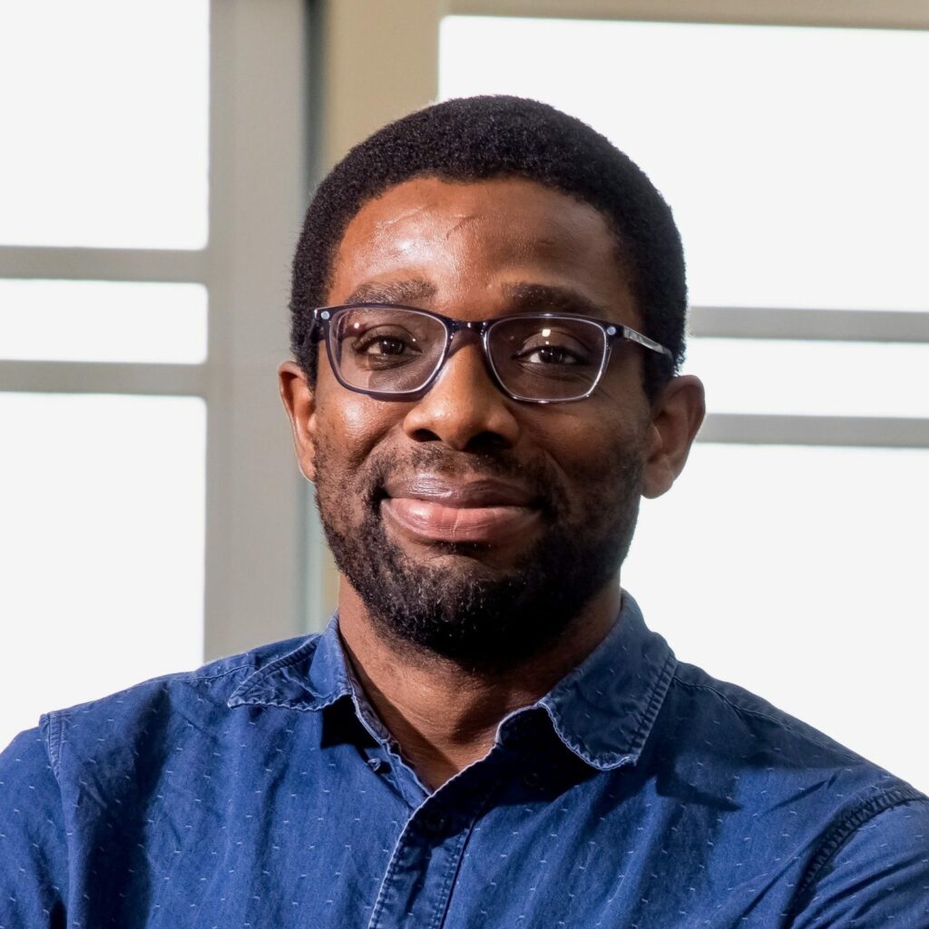 A man with short curly hair, a beard, and glasses is smiling slightly. He is wearing a blue button-up shirt and is standing indoors in front of large windows with bright light coming through.