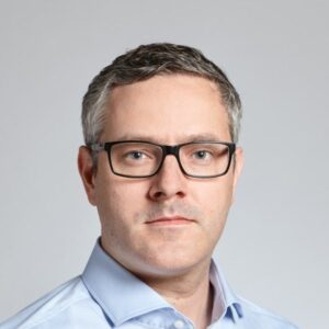 A man with short brown hair, wearing black rectangular glasses and a light blue collared shirt, looks directly at the camera with a neutral expression against a plain light grey background.