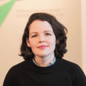 A woman with short dark hair and fair skin looks at the camera with a neutral expression. She is wearing a black jumper over a collared shirt, and the background is softly blurred with light colours.