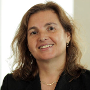 A woman with wavy brown hair, wearing a black blazer, a gold necklace, and small earrings, smiles at the camera against a softly lit, neutral background.