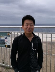 A person wearing a black shirt stands in front of a white fence with a beach and sea in the background under a cloudy sky.