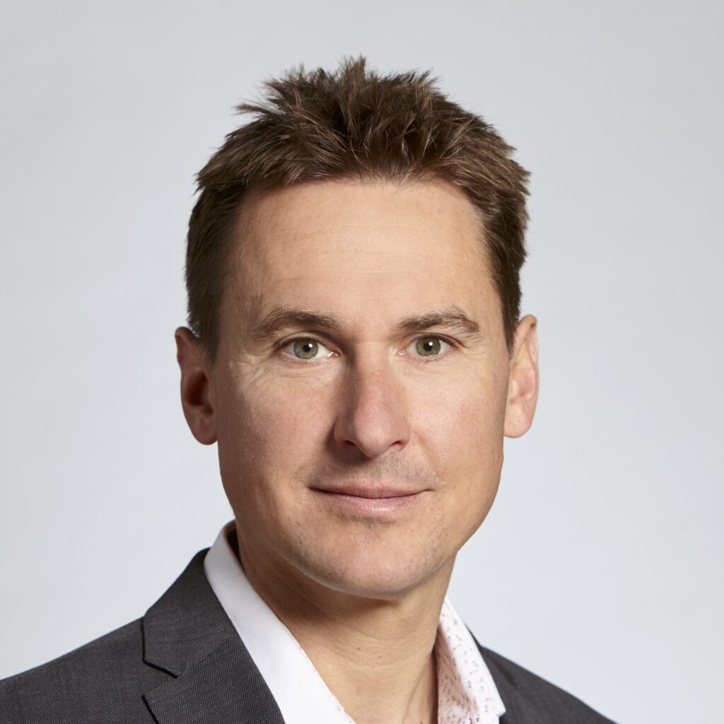 A man with short brown hair wearing a grey suit jacket and a white shirt, posing against a plain light grey background.
