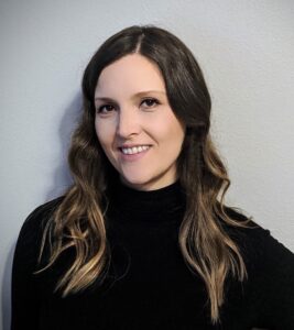 A woman with long, wavy brown hair and light highlights is smiling at the camera. She is wearing a black polo-neck jumper and is standing against a plain, light-coloured wall.