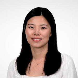 Portrait of a woman with straight black hair, wearing a white top and a delicate necklace, smiling softly in front of a plain white background.