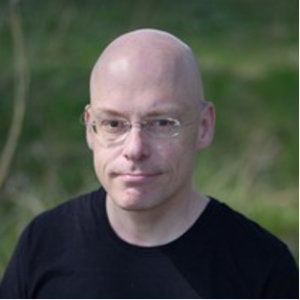 A bald person wearing glasses and a black shirt stands outdoors, with greenery blurred in the background.