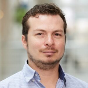 A man with short brown hair and a trimmed beard, wearing a light blue collared shirt, looks at the camera and smiles slightly. The background is softly blurred.