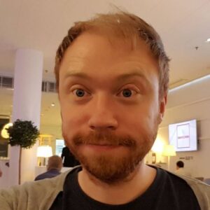 A man with light brown hair and a beard is smiling slightly while taking a selfie indoors. The background shows people sitting, modern decor, and warm lighting.