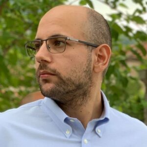 A man wearing glasses and a light blue button-up shirt looks to his left. He has short hair and a trimmed beard. Green leaves and blurred outdoor scenery are in the background.