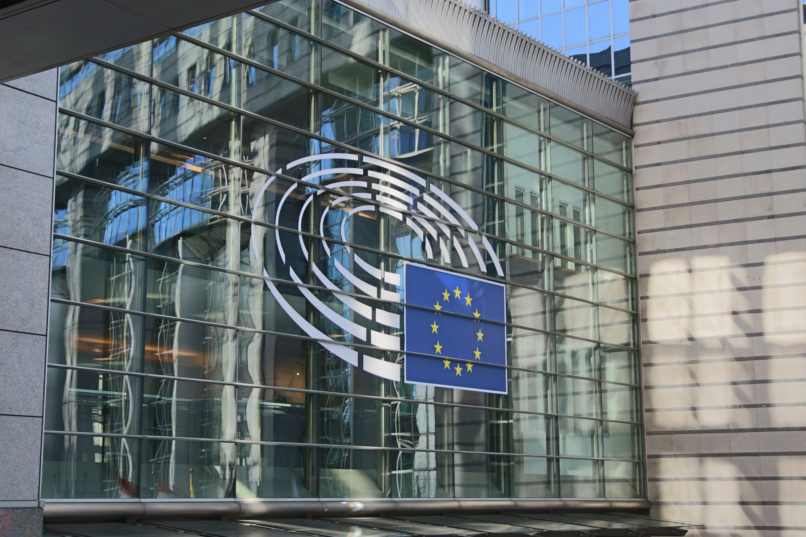 The photo shows the glass facade of a building with the European Union flag and the European Parliament logo displayed prominently on the windows. Reflections of nearby structures are visible on the glass.