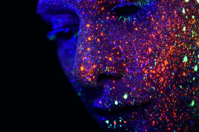 Close-up of a person's face covered in vibrant neon paint splatters, glowing under ultraviolet light in shades of purple, orange, and green, creating a striking, cosmic effect against a dark background.