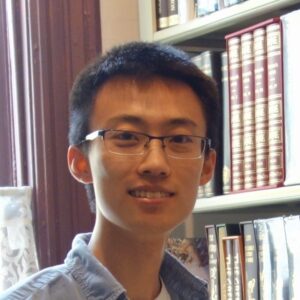 A young person with short black hair and glasses smiles in front of bookshelves filled with books in a library setting.