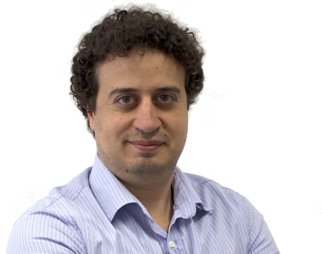 A man with curly dark hair and light skin, wearing a light blue striped shirt, smiles slightly whilst standing against a plain white background.