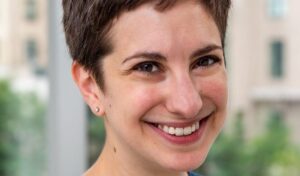 A woman with short brown hair smiles warmly at the camera. The background is softly blurred, showing green trees and beige buildings, suggesting an urban setting.
