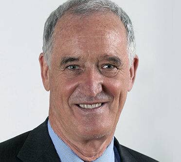 A smiling older man with short grey hair, wearing a blue dress shirt, striped tie, and dark suit jacket, standing in front of a plain light background.