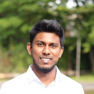 A man with short black hair and a trimmed beard smiles outdoors, wearing a white collared shirt. The background is blurred greenery and sunlight.