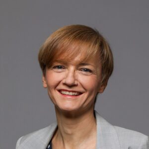 A person with short light brown hair is smiling and wearing a light grey blazer over a patterned top, posed against a plain grey background.