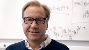 A middle-aged person with fair hair and glasses smiles gently, standing in front of a whiteboard covered with mathematical equations. They are wearing a dark jumper over a collared shirt.