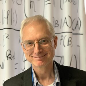 A smiling man with short, fair hair and glasses stands in front of a whiteboard covered with mathematical equations. He is wearing a dark blazer over a collared shirt.
