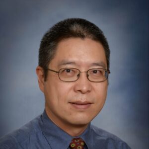 A man with short black hair and glasses wearing a blue collared shirt and a patterned red tie, posing in front of a grey-blue gradient background.