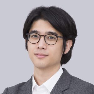 A person with medium-length black hair wearing round glasses, a white shirt, and a grey blazer, posing against a plain light grey background.