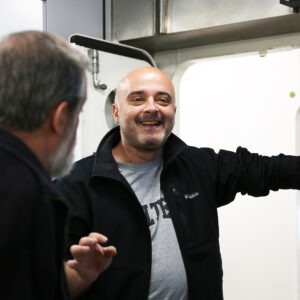 A man with a shaven head and a beard smiles whilst leaning against a doorway, talking to another man with grey hair and a beard inside a room with industrial equipment in the background.