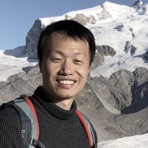 A man with short black hair, wearing a black polo neck jumper and a rucksack, smiles in front of a snowy mountain landscape under a clear blue sky.