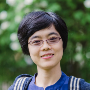 A person with short black hair and glasses smiles outdoors, wearing a blue top and a striped jumper. The background is blurred green foliage.