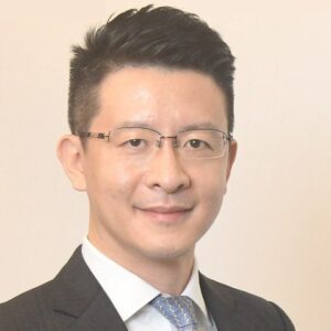 A man with short black hair, wearing glasses, a dark suit, a white shirt, and a blue patterned tie, smiles slightly in front of a plain light background.