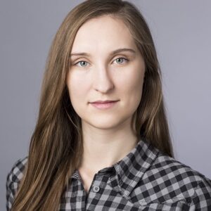 A woman with long, straight light brown hair and light eyes looks at the camera. She is wearing a black and white chequered shirt and is posed in front of a plain, light grey background.