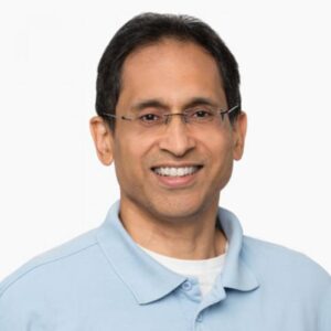 A smiling man with short dark hair, wearing glasses and a light blue collared shirt over a white T-shirt, poses against a plain white background.
