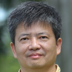 A middle-aged man with short, grey and black hair smiles slightly whilst standing outdoors, with a blurred background of green foliage and tree trunks.