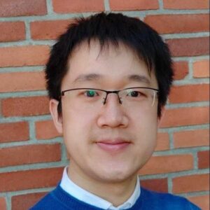 A person with short black hair and glasses, wearing a blue jumper over a white collared shirt, is smiling slightly whilst standing in front of a brick wall.