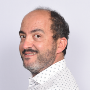 A man with short dark hair and a receding hairline, wearing a white shirt with small grey dots, is smiling and looking slightly over his shoulder against a plain light background.