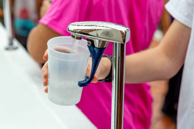 A person fills a clear plastic cup with water from a metal dispenser, wearing a white shirt and black wristband, with another person in a pink shirt in the background.