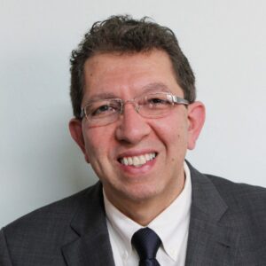 A man wearing glasses, a grey suit, white shirt, and dark tie smiles at the camera against a plain light-coloured background.