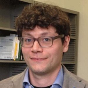 A person with short, curly brown hair and glasses is smiling slightly, wearing a blue shirt and grey blazer, standing in front of shelves with books and files.
