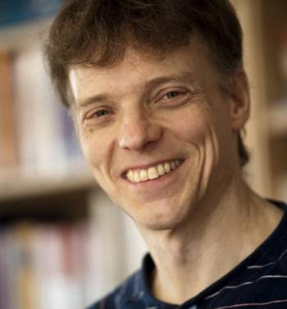 A man with short brown hair smiles at the camera, wearing a dark striped shirt. The background is softly blurred with bookshelves visible, suggesting an indoor setting.