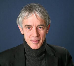 A middle-aged man with grey hair wearing a black polo-neck jumper and black blazer, smiling softly in front of a plain dark blue background.