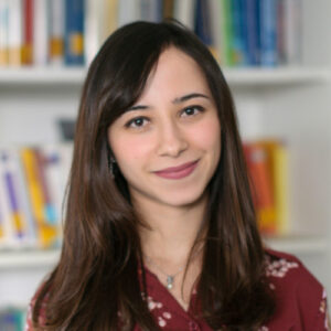 A young woman with long brown hair and a maroon blouse smiles in front of a blurred bookcase filled with colourful books.