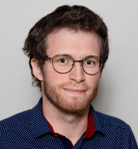 A young man with short brown hair, a beard, and round glasses smiles slightly. He is wearing a navy blue shirt with white dots and a red collar, and stands against a plain light grey background.