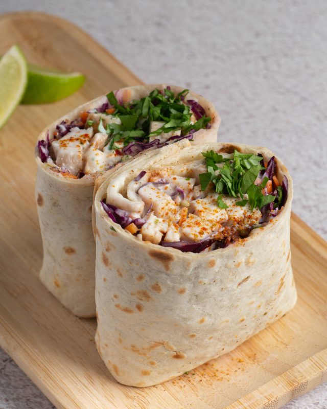 Two halves of a wrap stuffed with grilled chicken, shredded red cabbage, and fresh herbs are placed on a wooden tray. Two lime wedges sit in the background. The wraps are sprinkled with spices and chopped parsley.