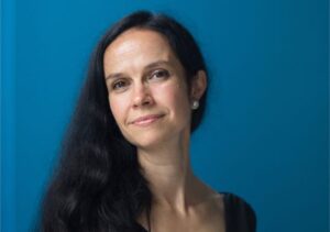 A woman with long black hair and pearl earrings smiles gently at the camera against a solid blue background. She is wearing a black top.