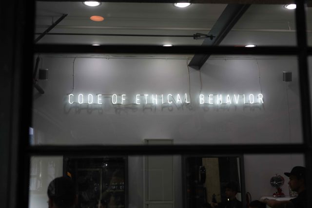 A neon sign on a white wall reads CODE OF ETHICAL BEHAVIOUR in uppercase letters, viewed through a window with black frames. There are a few people partially visible inside.