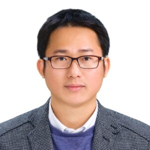 A man with short black hair and glasses, wearing a grey checked blazer over a white collared shirt and a blue jumper, poses against a plain white background.