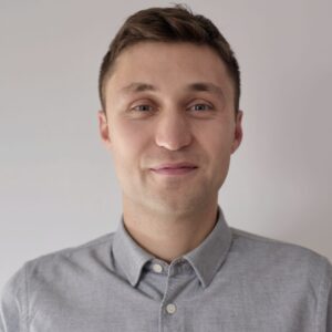 A young man with short brown hair and blue eyes is smiling softly, wearing a light grey button-up shirt, standing against a plain light-coloured background.