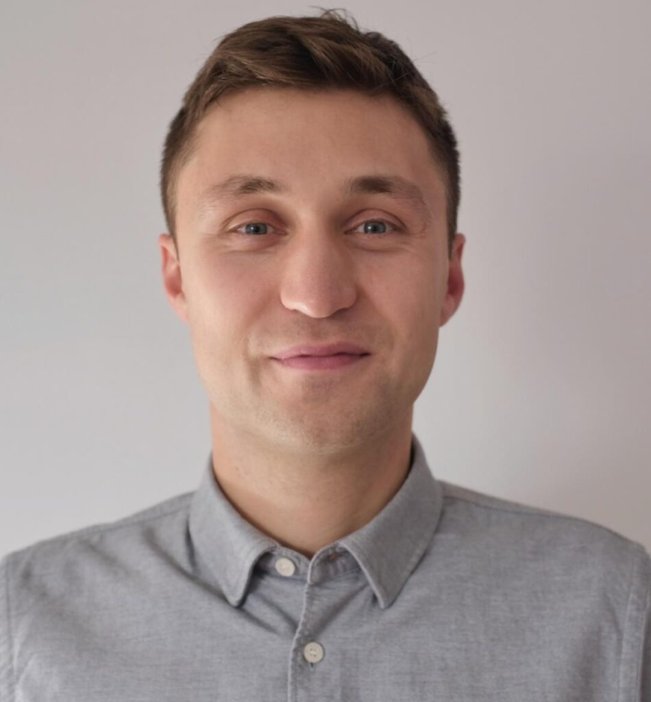 A man with short brown hair, wearing a light grey button-up shirt, stands in front of a plain light-coloured background, smiling softly at the camera.