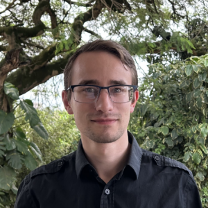 A person with short brown hair and glasses wearing a dark button-up shirt stands outdoors in front of lush green foliage and trees.